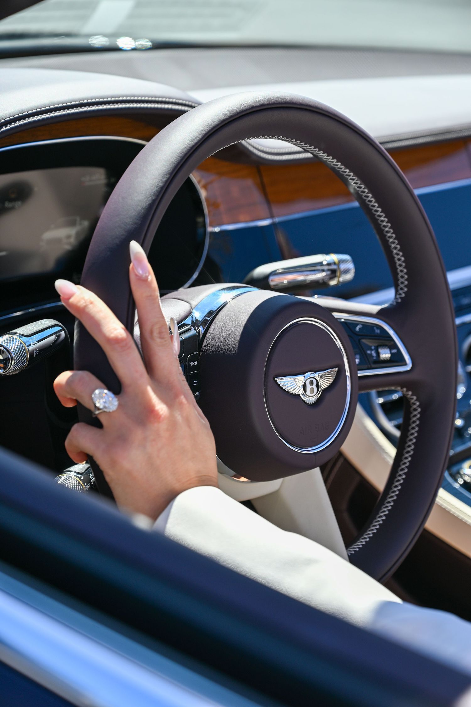 A manicured hand with long nails rests on a luxury Bentley steering wheel with wood trim and chrome accents.