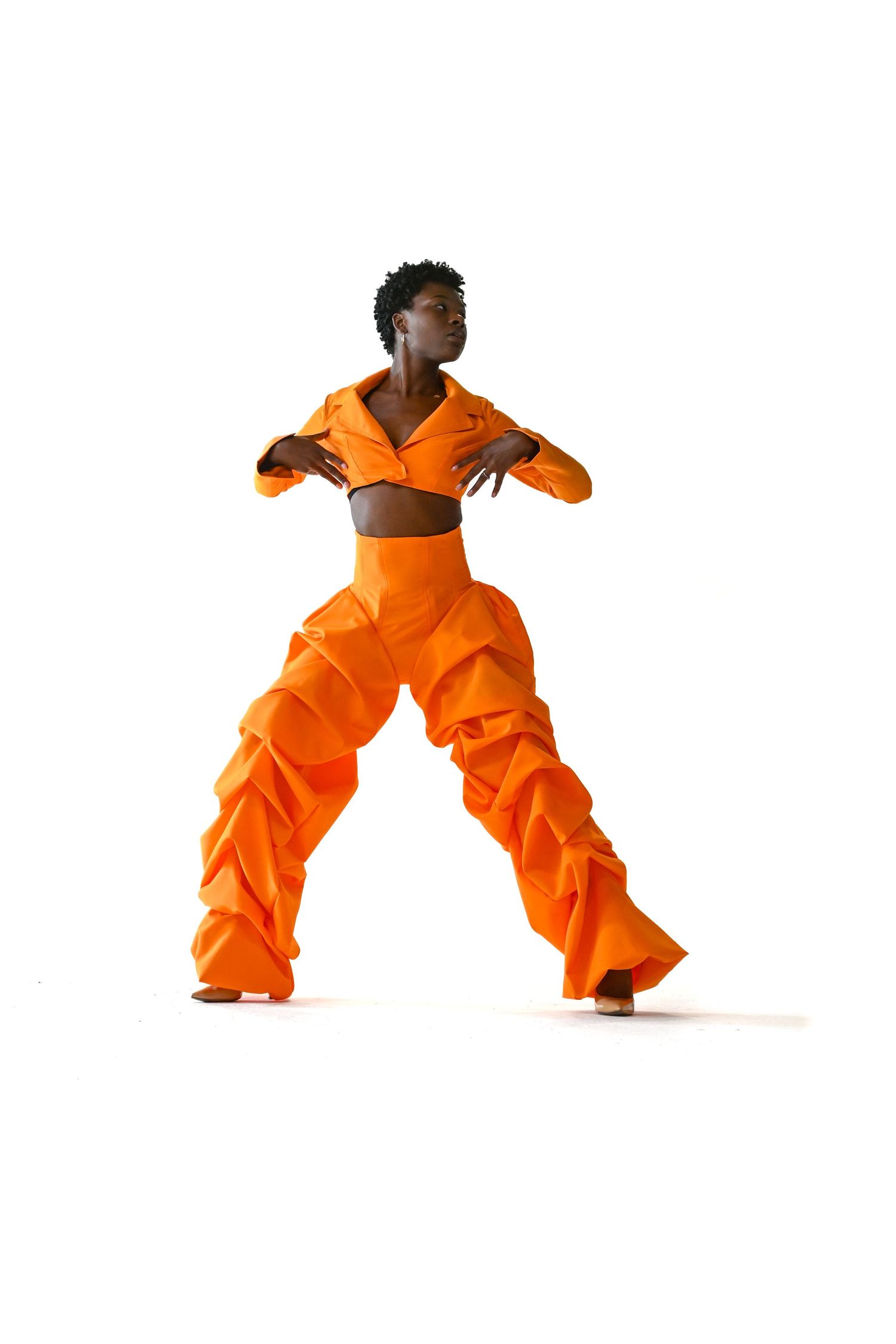 Dancer in vibrant orange two-piece outfit with ruched wide-leg pants strikes dynamic pose against white background.
