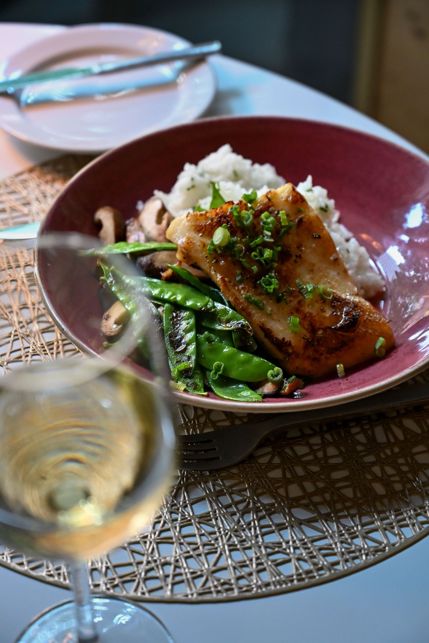 Asian-inspired glazed fish dish served with white rice and vegetables alongside a glass of white wine in burgundy bowl.