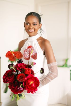 Bride in sleek white halter dress poses with stunning red floral bouquet.