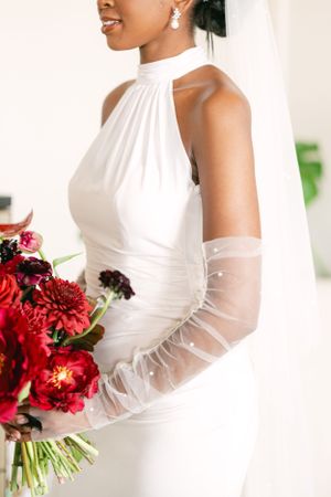 Elegant bride in halter neck dress holds vibrant red bouquet.