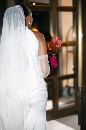 Bride in elegant white dress holds red flowers while standing near a doorway.