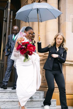 A bride in a white dress with red roses is assisted down stairs under a grey umbrella on a rainy day.