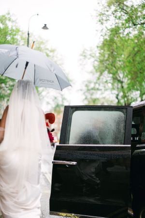 A white wedding dress and veil visible under a grey umbrella as someone enters a black car.