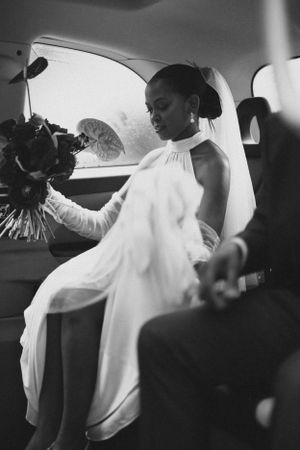 Bride in flowing white halter dress seated in wedding car with bouquet.