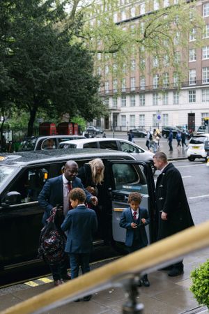 A group gathers around a black car on a rainy city street with historic buildings in the background.
