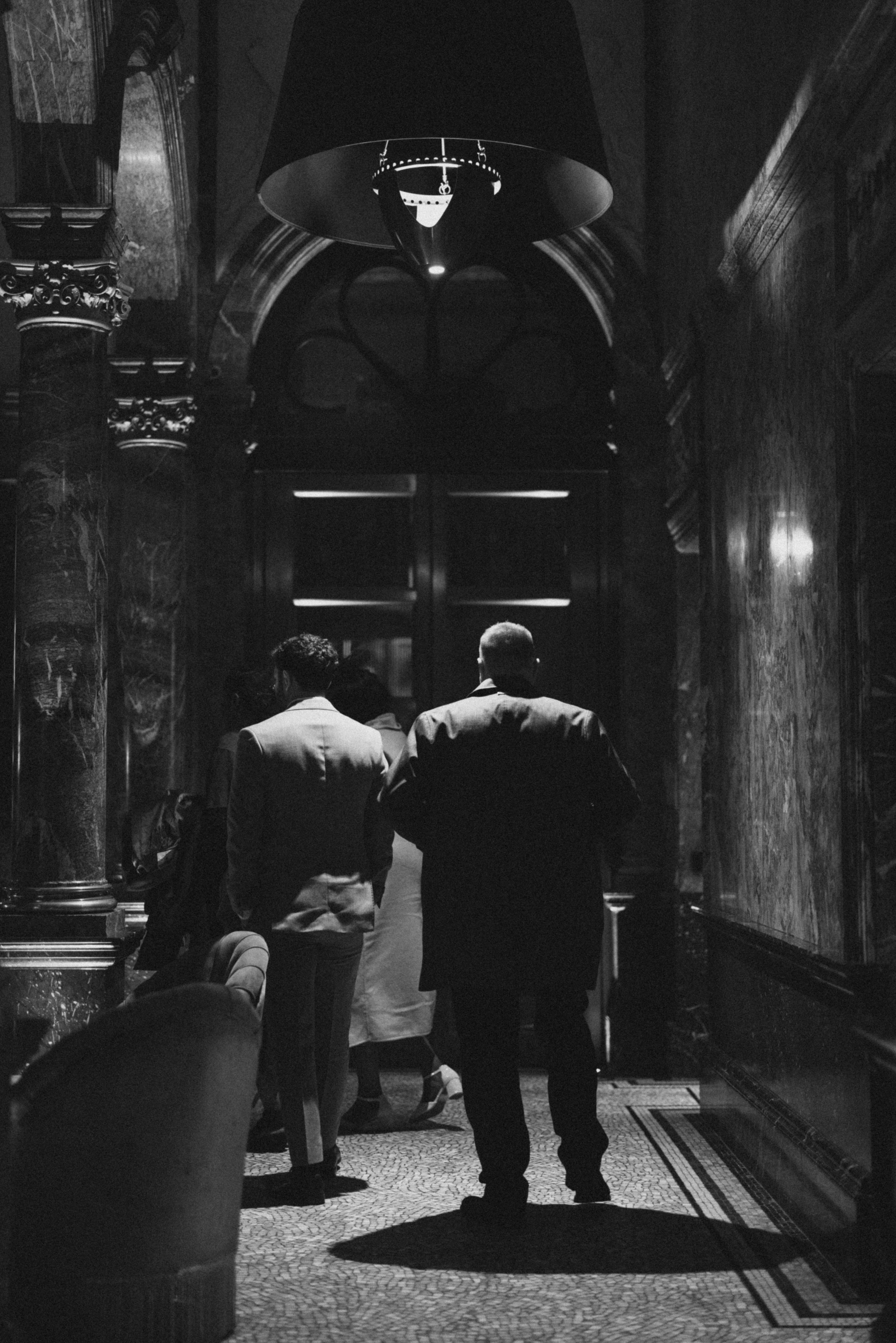 Shadowy figures stand beneath an ornate arched doorway in dramatic black and white lighting.