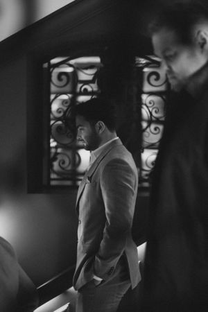 Men in formal attire stand near decorative wrought iron windows in moody black and white photography.