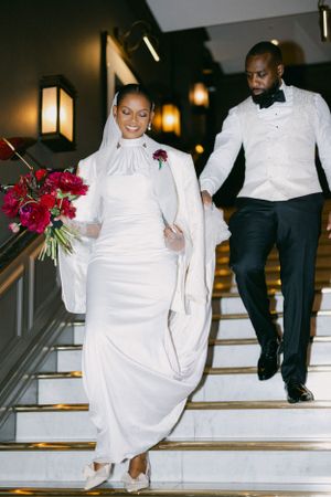 A newlywed couple descending white stairs, bride in elegant white dress with red bouquet.