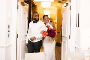 Newlyweds walking through a brightly lit hotel hallway with red floral bouquet.