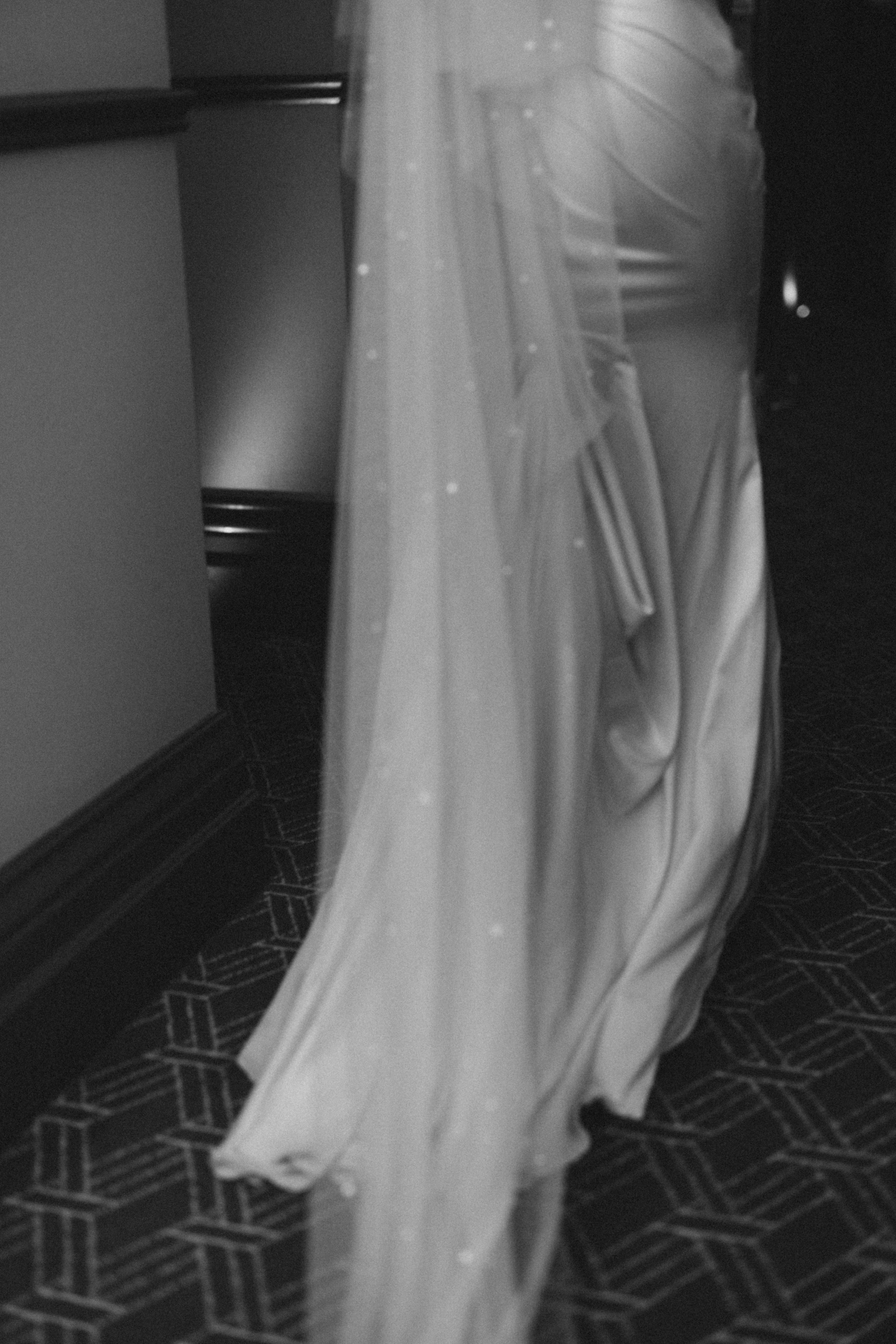 Black and white detail shot of flowing wedding dress and veil against geometric carpet.