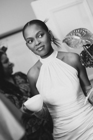 A stylish bride in a white halter dress poses with a drink at her wedding reception.