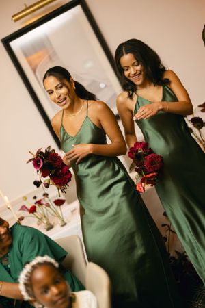 Bridesmaids in sage green dresses hold burgundy bouquets while laughing.