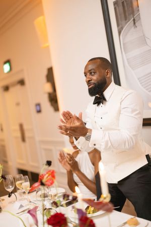 Groom in white tuxedo jacket applauds during wedding reception dinner.