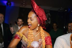 A celebrant wearing a red headwrap and orange beaded necklace dances joyfully at a formal event.