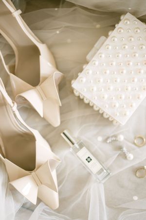 Elegant white bridal shoes, perfume bottle and pearl clutch arranged on tulle fabric.