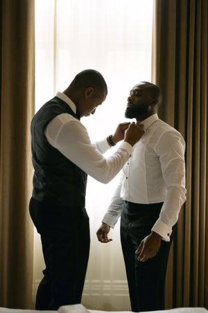 Silhouetted figures adjusting formal attire near a window.