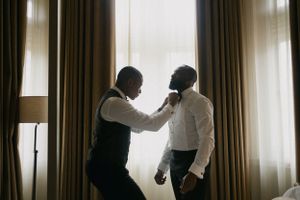 Two people silhouetted against window light during formal dressing preparation.