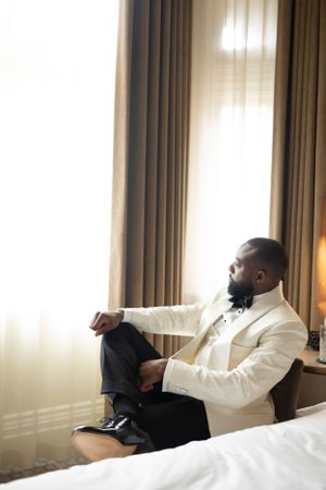 Person in white tuxedo jacket sitting on edge of bed near window.