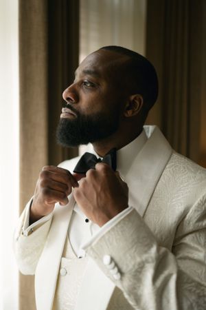 A man in a white tuxedo jacket and bow tie adjusts his collar while looking to the side in elegant lighting.