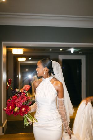 Bride in elegant white halter dress holding red bouquet in hotel hallway.