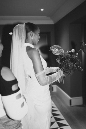 A bride in a sleek white dress and long gloves holds flowers while smiling in profile.