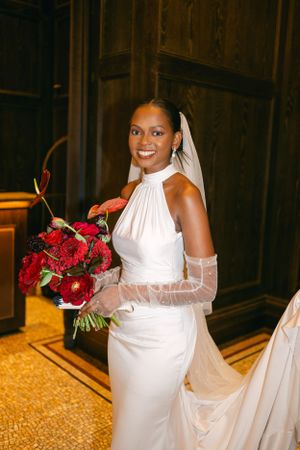 Elegant bride holding red bouquet in white halter gown with sheer sleeves.