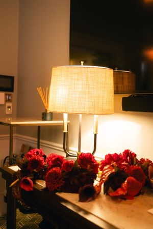 Table lamp glows warmly beside red flower arrangement in evening setting.