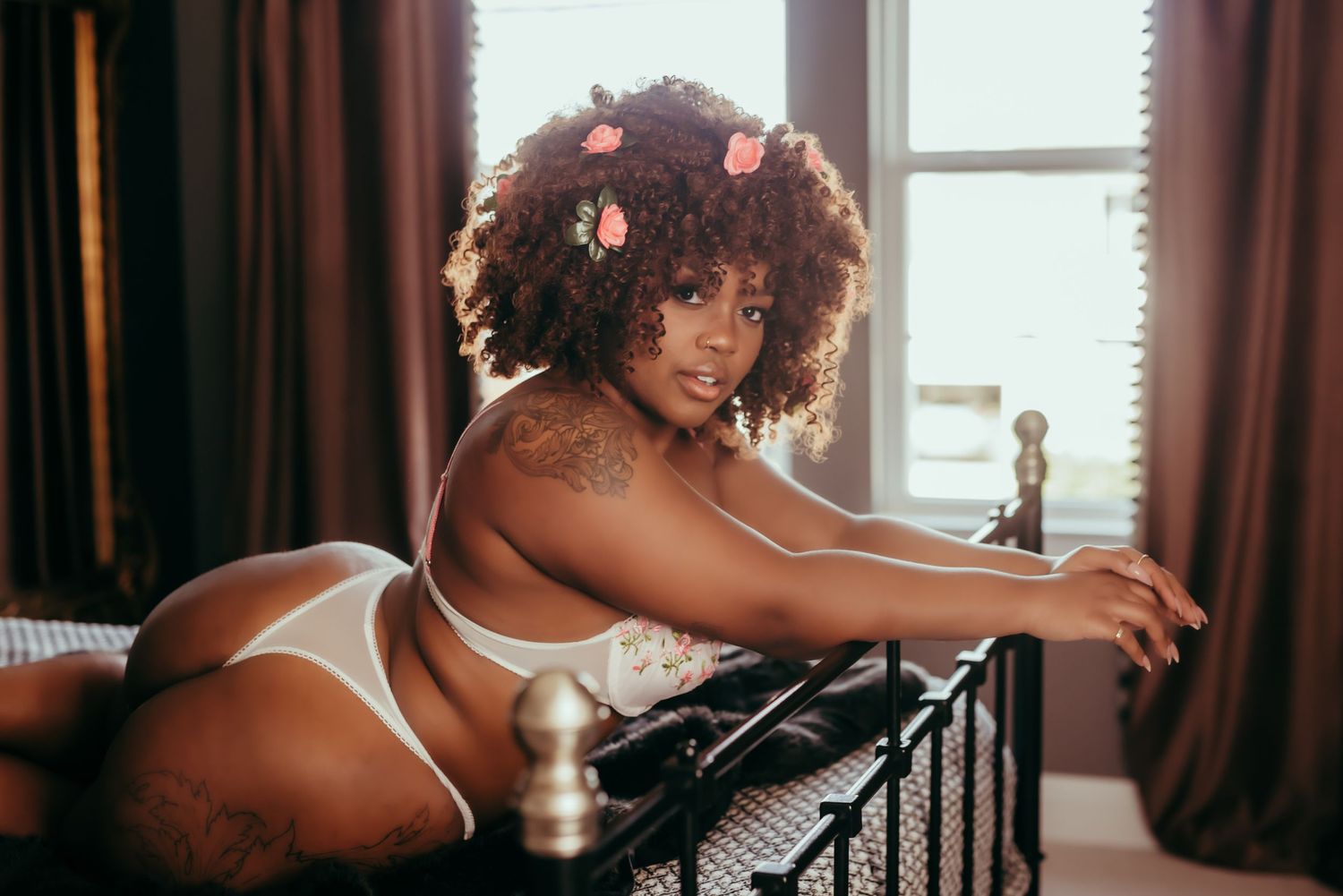 A young woman in white lingerie poses seductively on an ornate iron bed frame by a window with brown curtains.