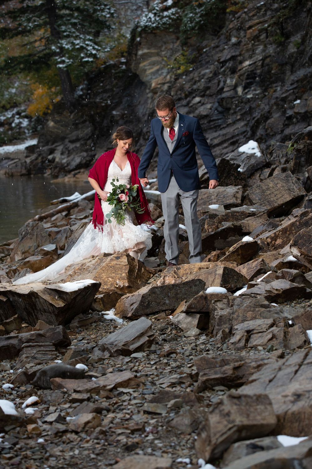 A couple in wedding attire carefully navigates rocky terrain near a cliffside during their outdoor mountain photo session.