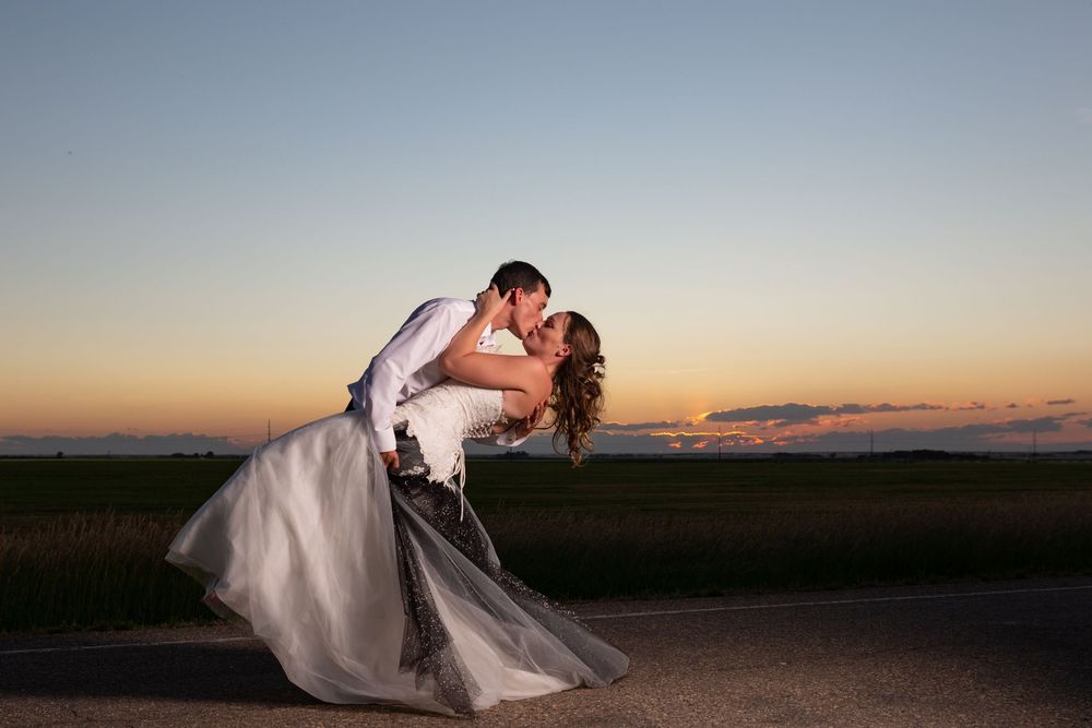 A romantic couple in wedding attire share an intimate dip pose against a stunning sunset sky.