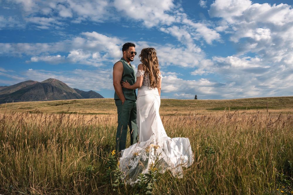 A couple stands together in a golden field with mountains and blue skies in the background.