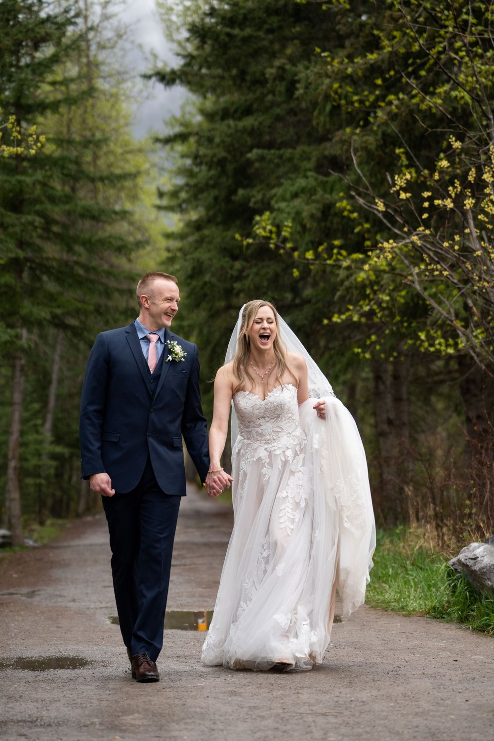 A couple walks down a wooded path in wedding attire, with the bride wearing a flowing white lace dress and veil.