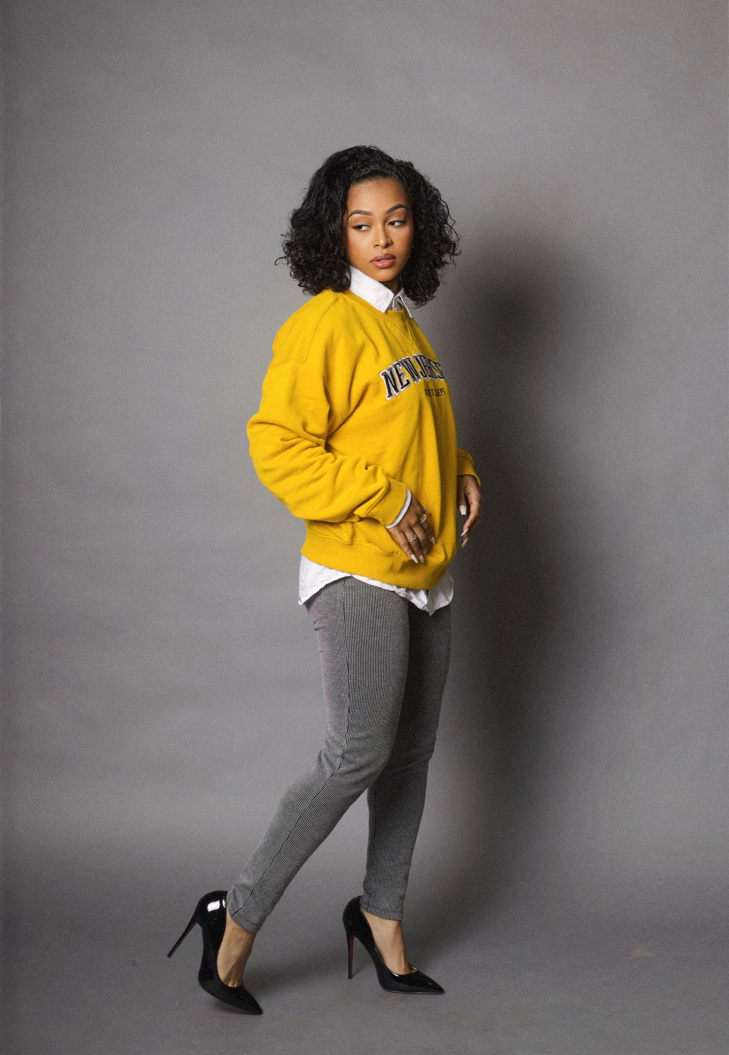 Fashion model poses in a bright yellow sweater and striped pants against a gray studio backdrop.