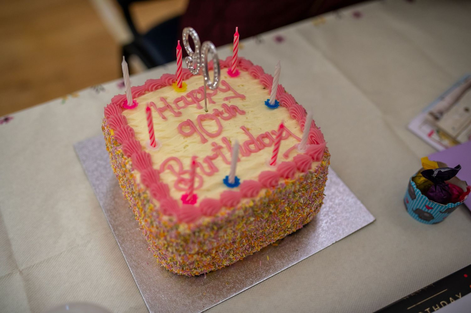 Pink birthday cake with sprinkles and colorful candles on white surface.