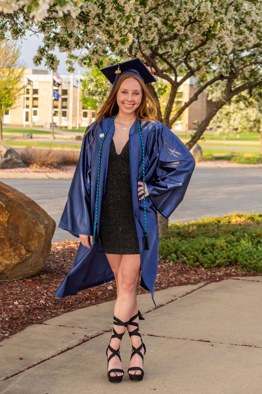 Graduate in blue cap and gown poses near campus building on sunny day.