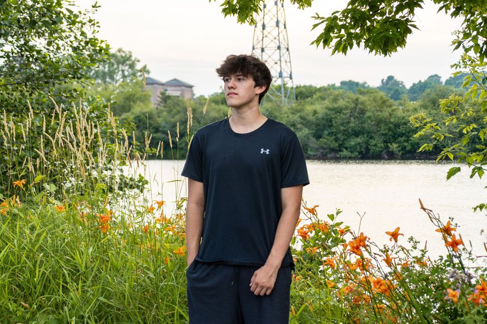 Person in an Under Armour t-shirt stands near a lake with orange wildflowers and lush greenery in the background.