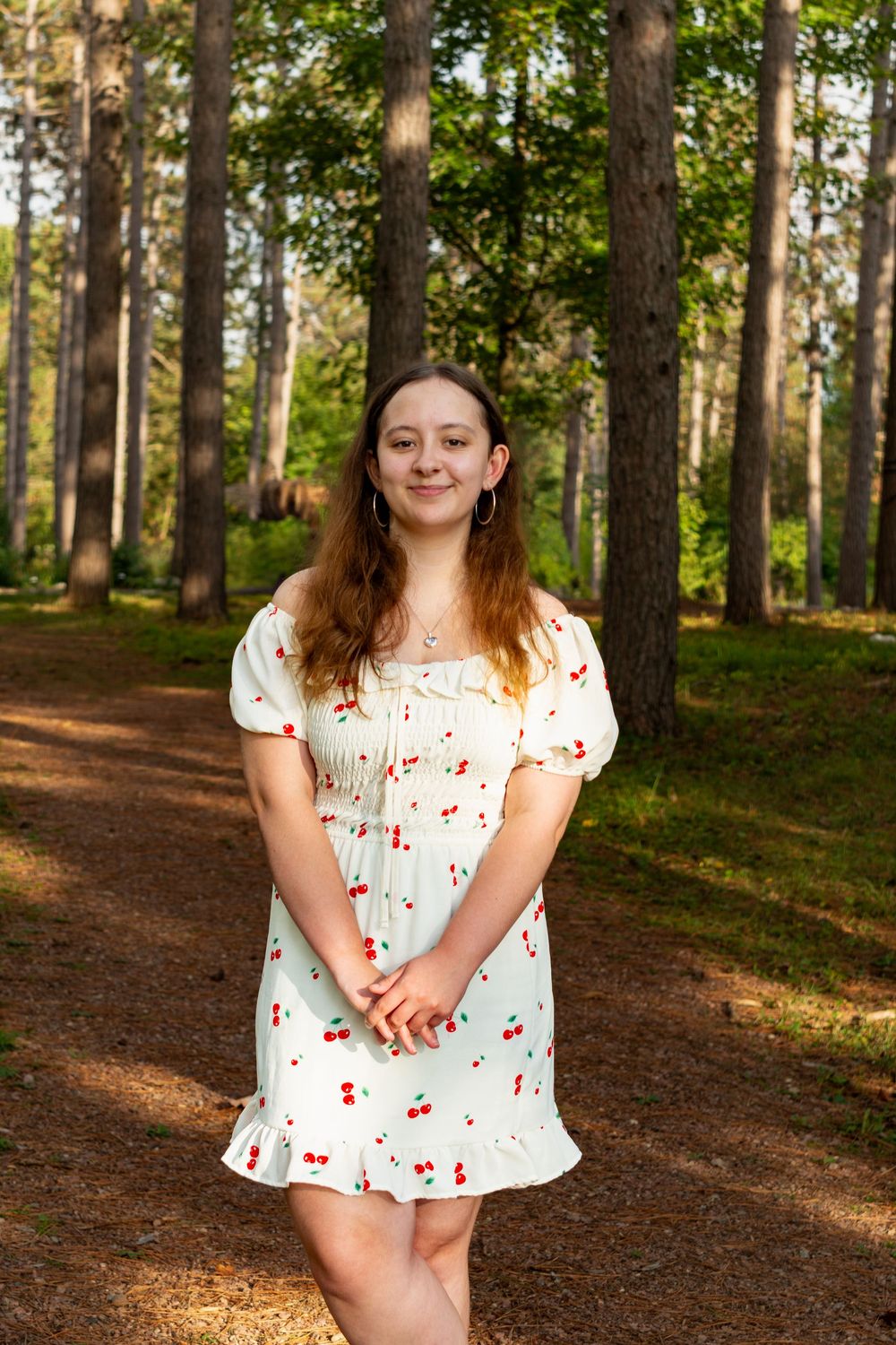 A person wearing a white cherry-print dress poses in a sunlit forest setting.