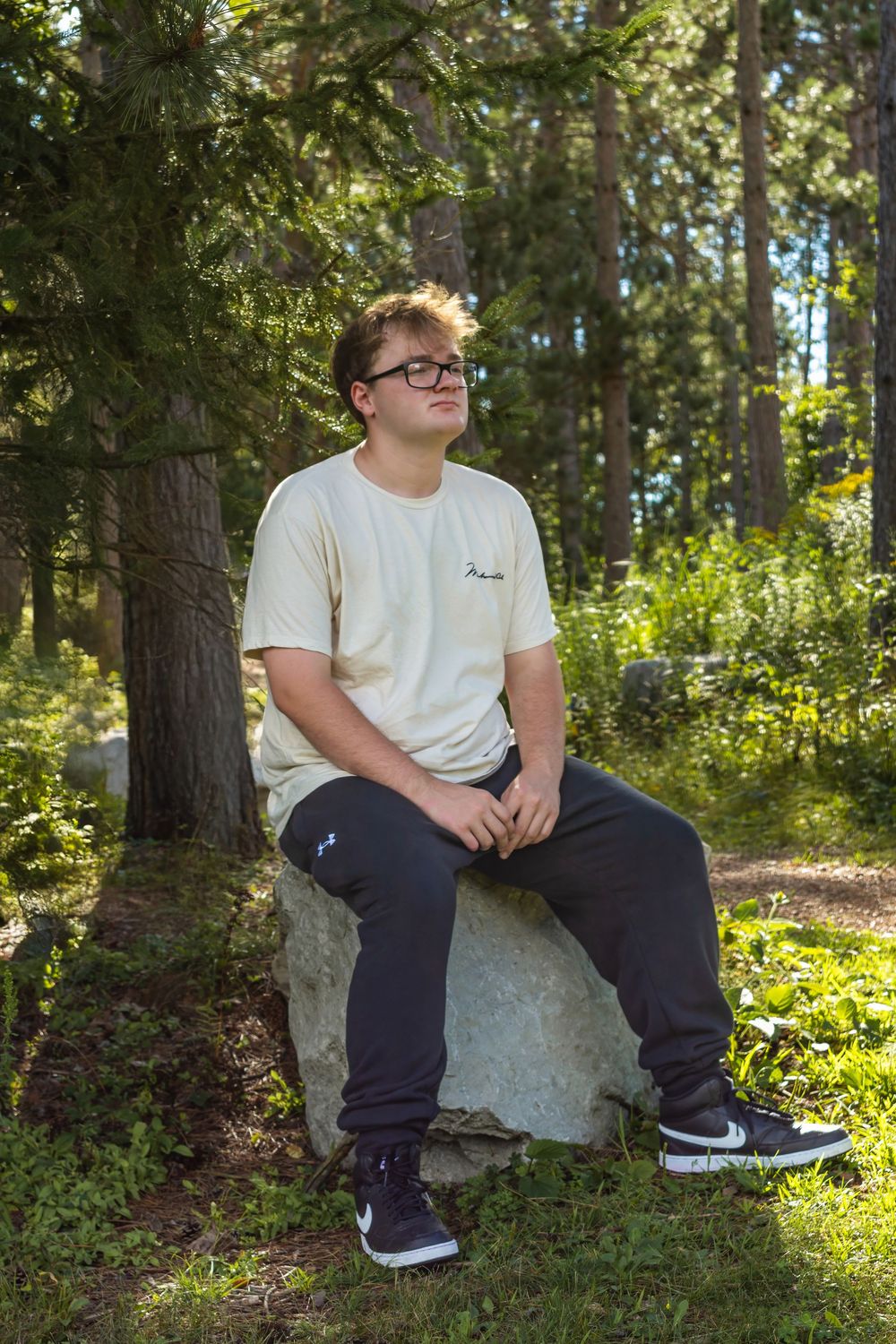 Person in white t-shirt and navy pants sits on rock in sunlit forest clearing.