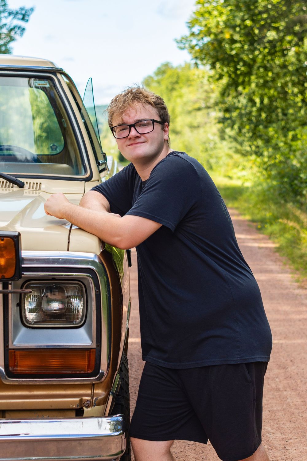 A vintage beige car parked on a dirt road surrounded by green trees and natural scenery.