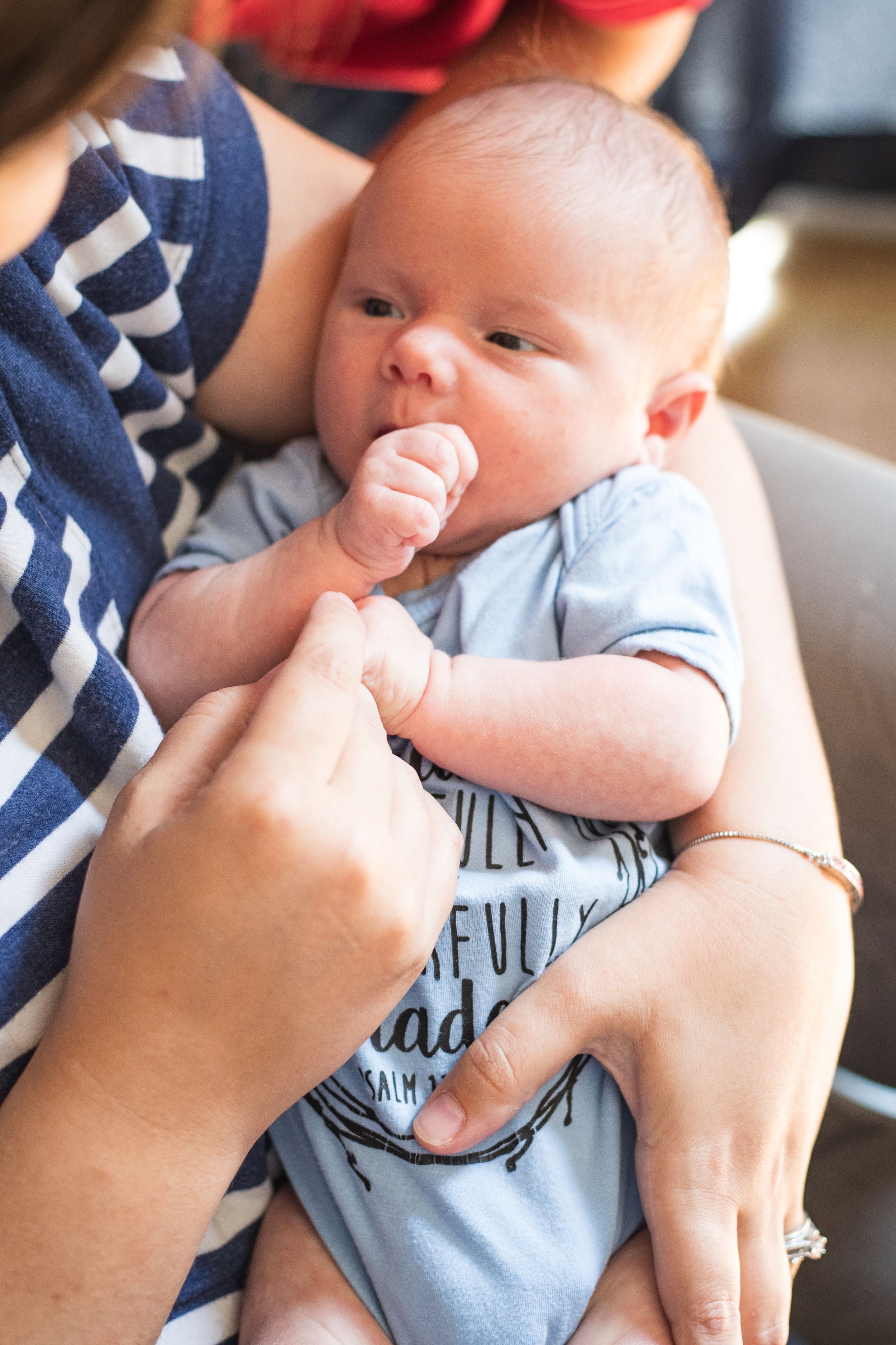 A newborn baby in a blue onesie grasps an adult finger while being cradled in loving arms.