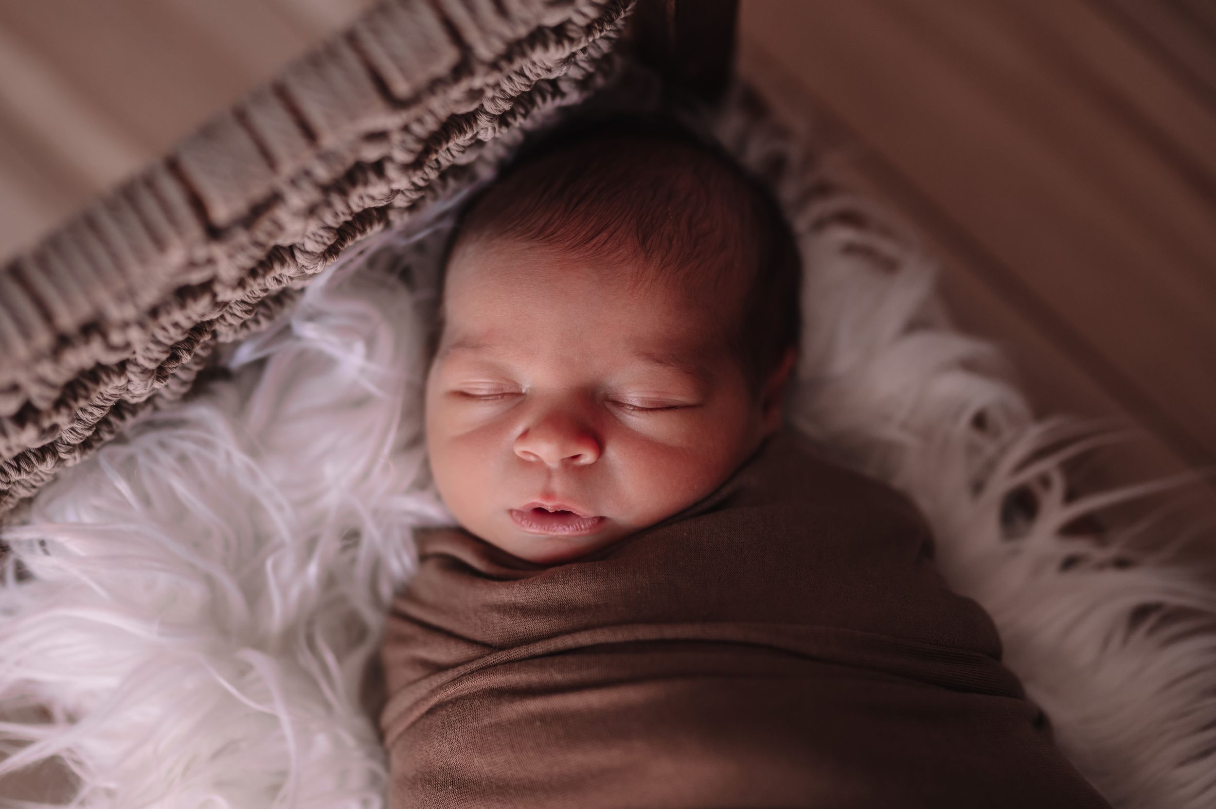Sleeping newborn wrapped in brown swaddle on white furry backdrop.
