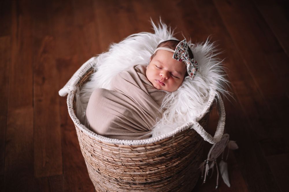 A peaceful newborn baby sleeps in a woven basket on soft white fur.
