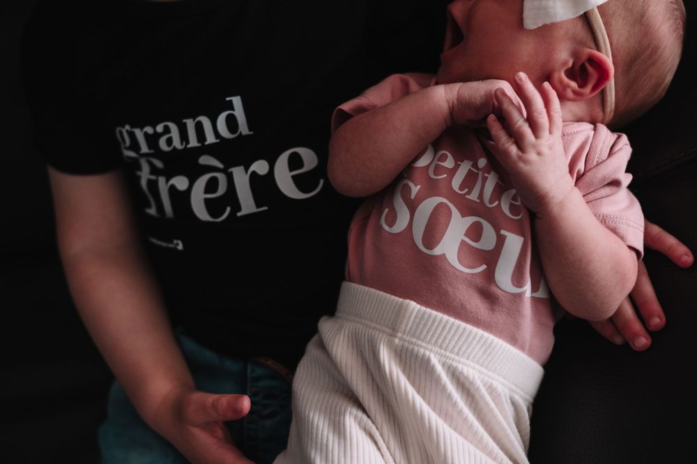 A newborn baby wearing pink being held while someone wears a 'grand frere' shirt.