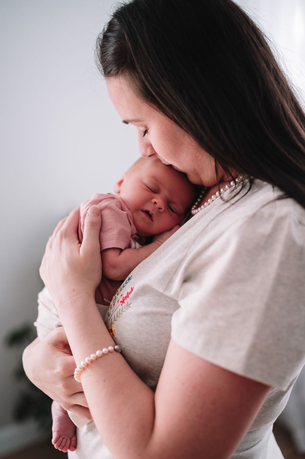 A tender moment as a parent in white holds and kisses their sleeping newborn baby against a light background.
