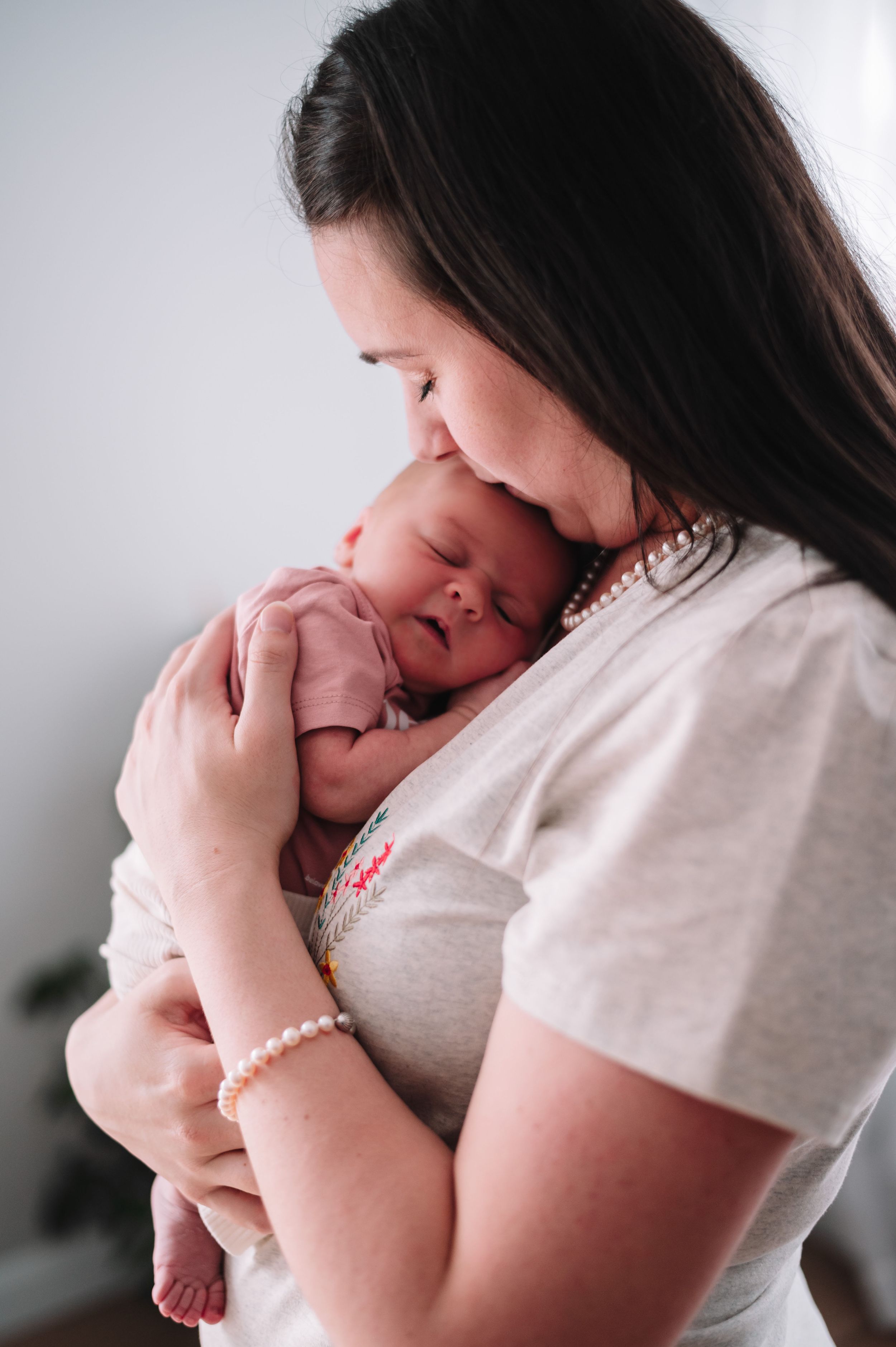 A tender moment as a parent in white holds and kisses their sleeping newborn baby against a light background.