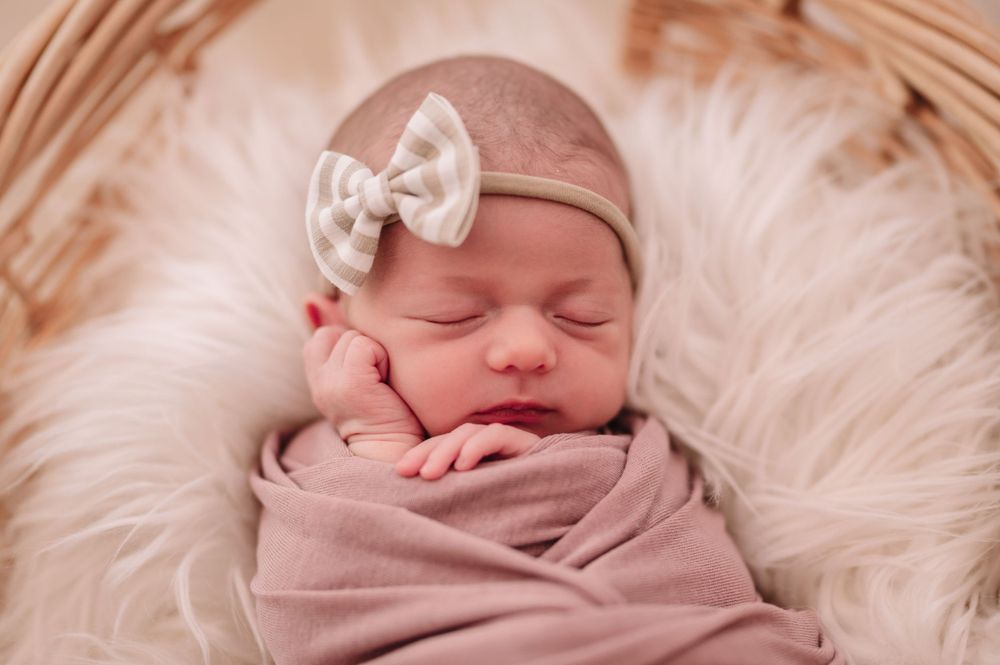 Sleeping newborn wrapped in pink fabric wearing a bow headband on fluffy white fur.