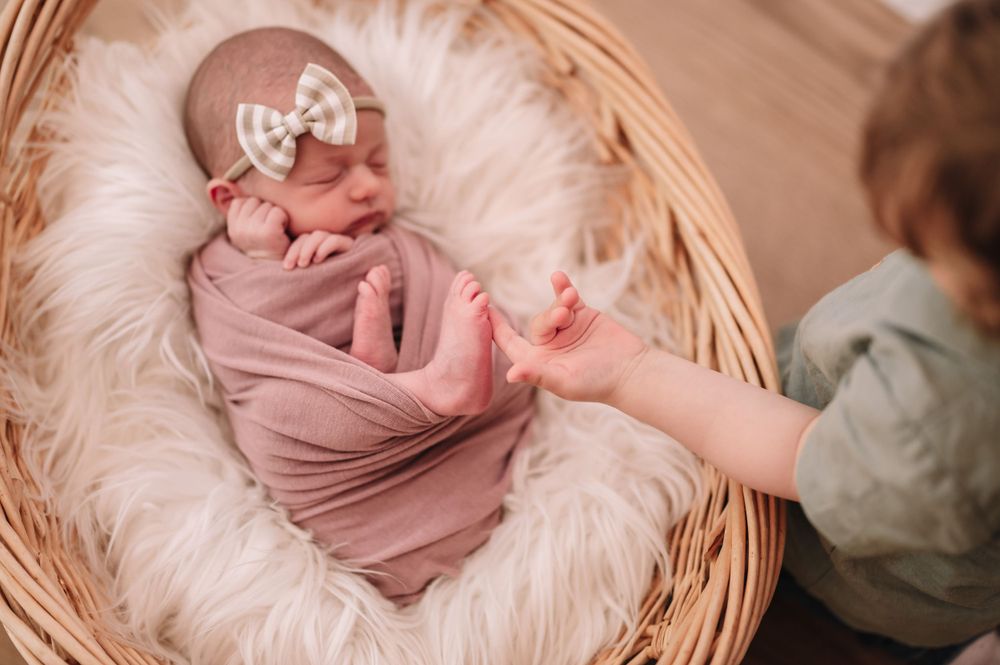Baby wrapped in pink with bow headband sleeping in basket while sibling reaches to touch.