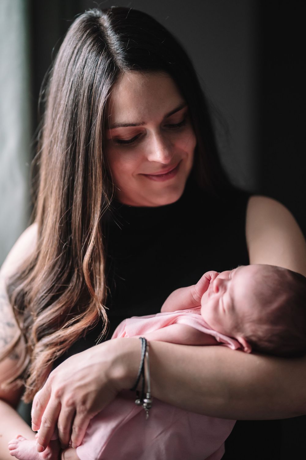 Parent in black top gazes lovingly at sleeping newborn baby during intimate portrait session.