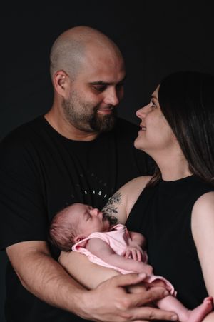 Parents in dark clothing share loving glance while holding their sleeping newborn.